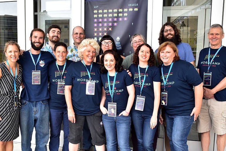 Group of WordCamp organizers stand smiling in front of 2018 banner