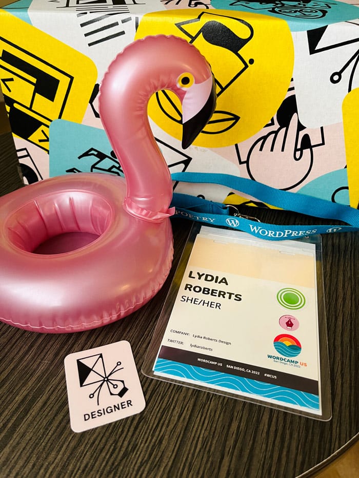 Lydia’s WordCamp name badge lays on a table next to swag items like a small pink inflatable flamingo, designer sticker, and a patterned box full of Godaddy items.