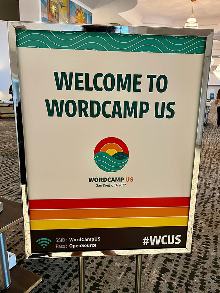 A colorful printed sign reads "Welcome to WordCamp US" inside a carpeted hotel lobby